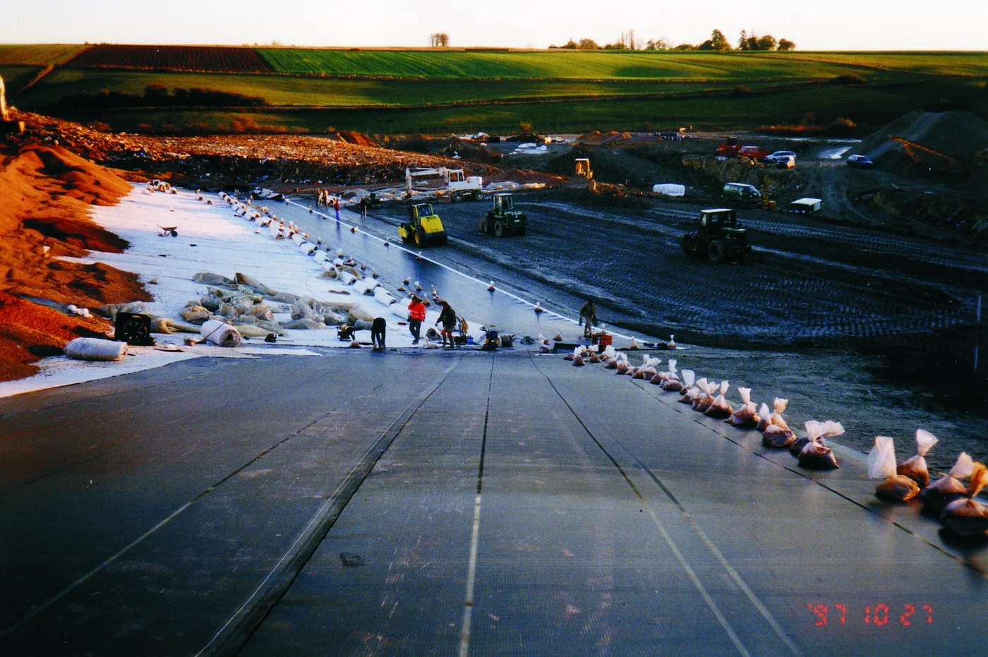 Workers unrolling black HDPE geomembrane sheets across a compacted base liner at a landfill construction site, with welded panel seams and anchor trench