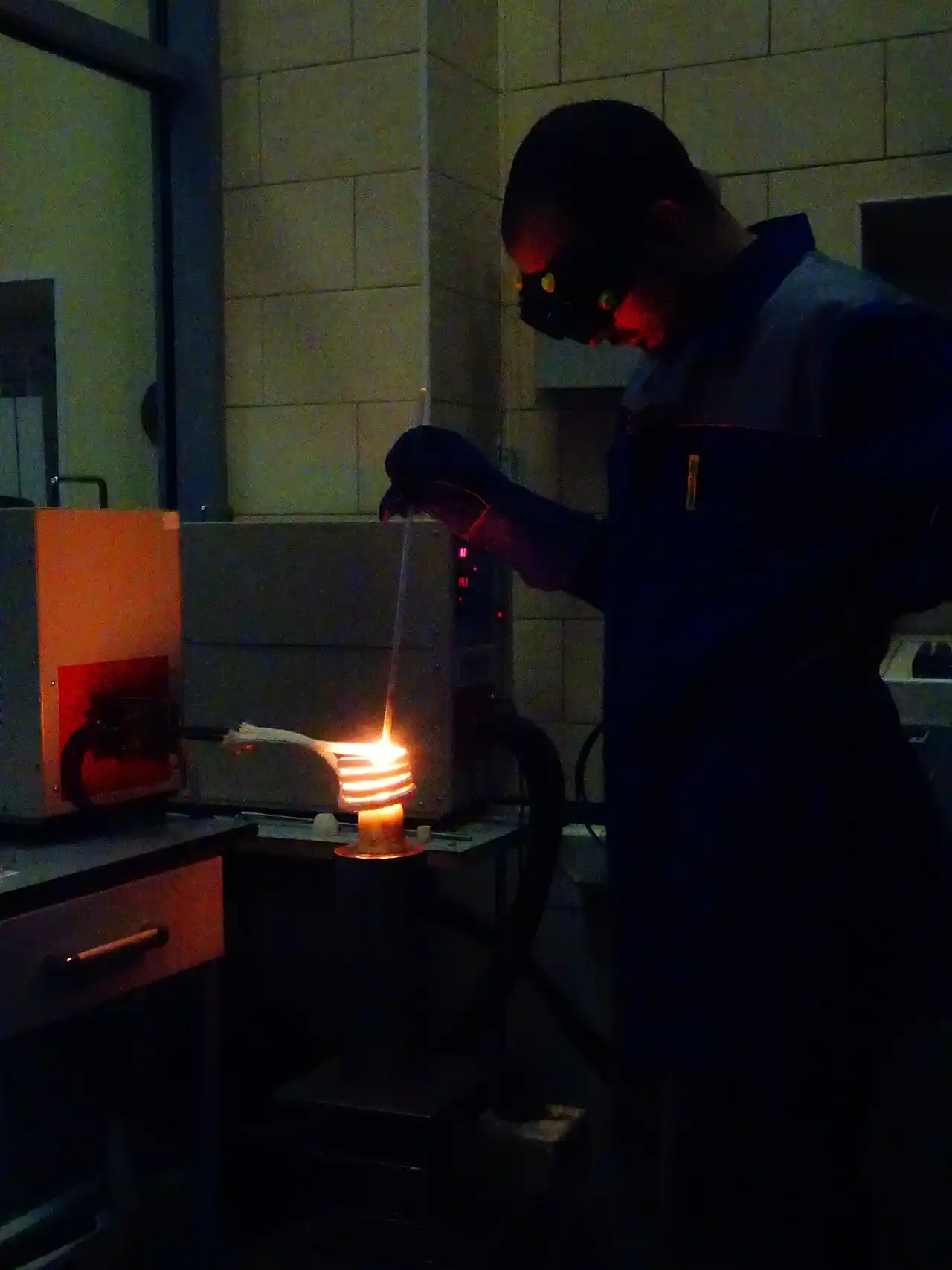 Operator melting metal in a small induction furnace: a glowing water-cooled copper induction coil heats a crucible while the medium-frequency power supply runs alongside