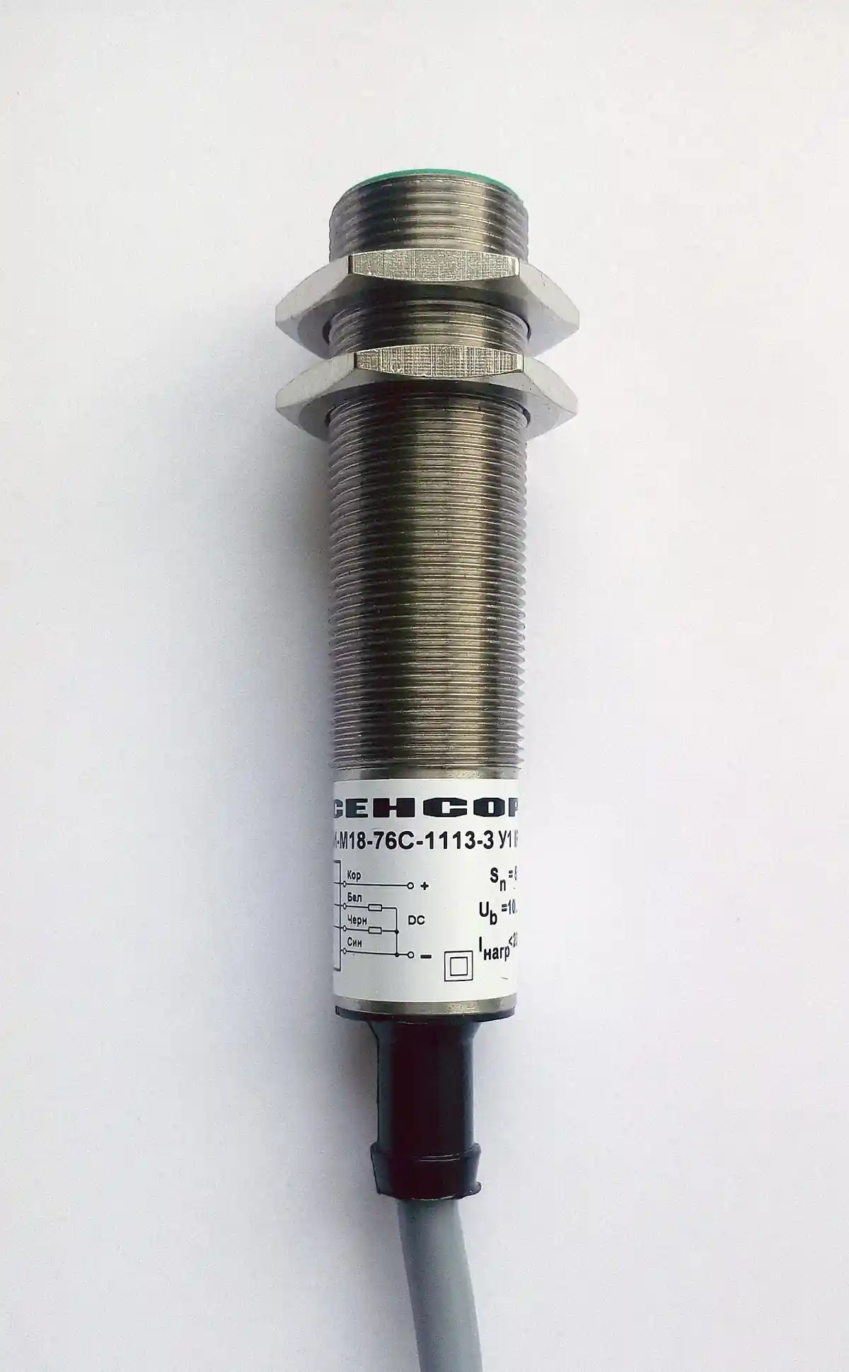 M18 threaded cylindrical inductive proximity sensor with nickel-plated metal barrel, hex mounting nuts, green active-face ring, wiring-diagram label and integral cable