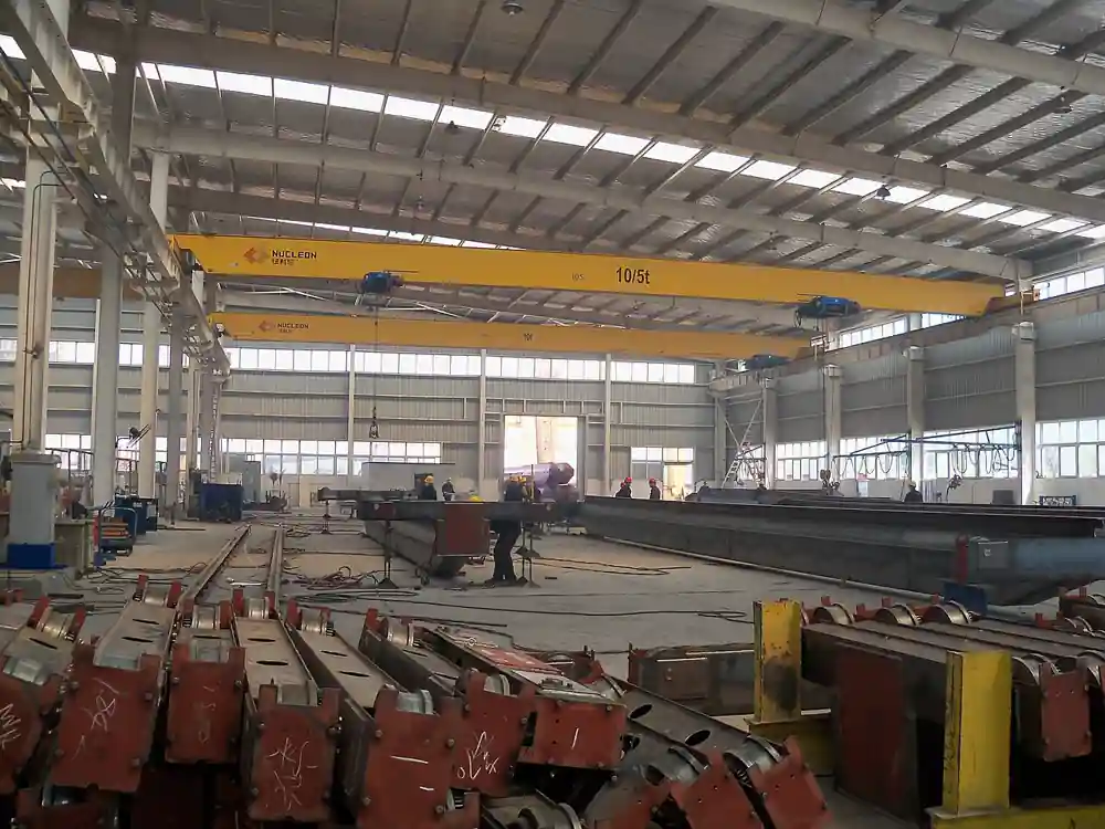 Two yellow LD-model single girder overhead travelling cranes spanning a steel-fabrication workshop bay, each with a single bridge box girder, a 10/5 ton hoist trolley, and end trucks riding the elevated runway