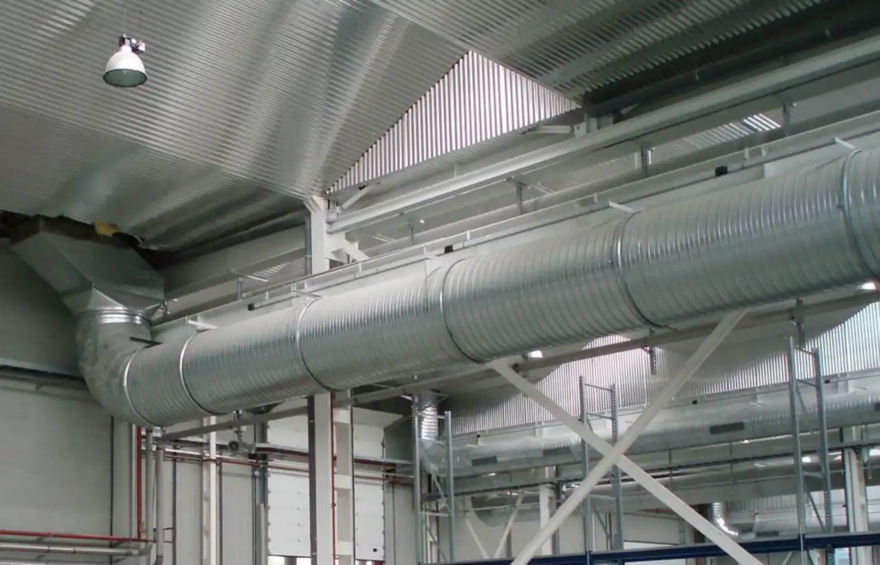 Round galvanized spiral duct with visible helical lockseam running across an industrial warehouse ceiling, with branch fittings and a transition