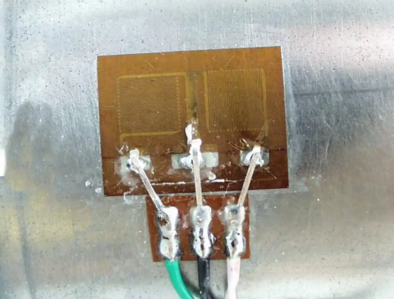 Bonded metallic foil strain gauge with two photo-etched grids on an amber polyimide backing, bonded to a metal surface with three soldered lead wires