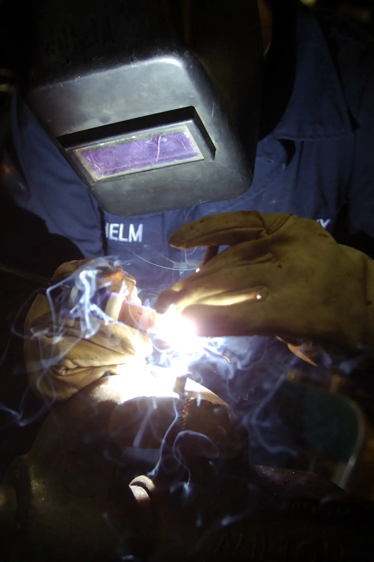 A welder in a protective helmet and gloves welds a threaded metal stud onto a workpiece, with a bright welding arc and smoke at the joint