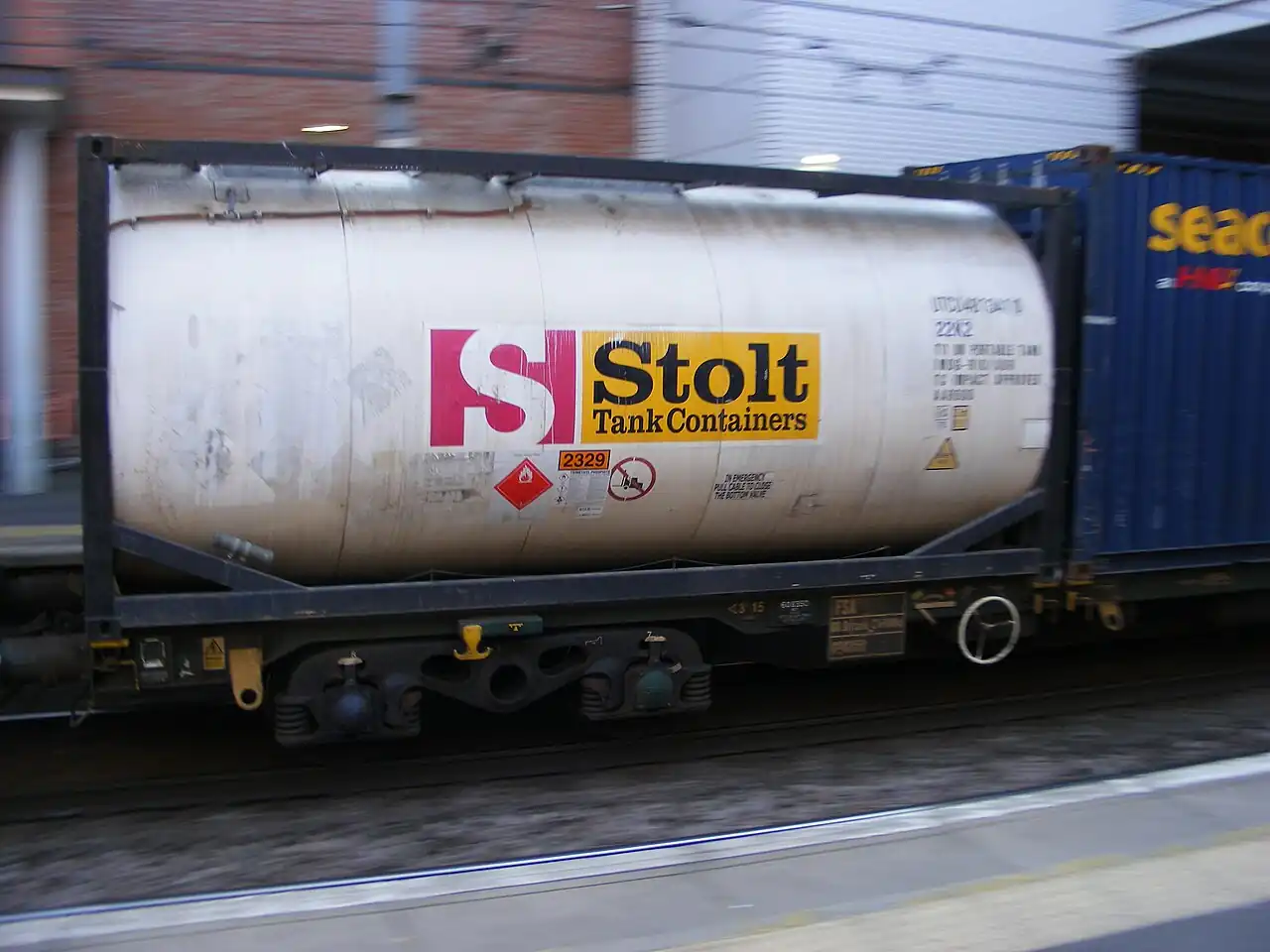 A Stolt 22K2 20-foot ISO tank container, a cylindrical stainless-steel pressure vessel mounted inside a standard ISO freight-container frame, on a rail wagon with UN portable tank markings and a hazmat placard