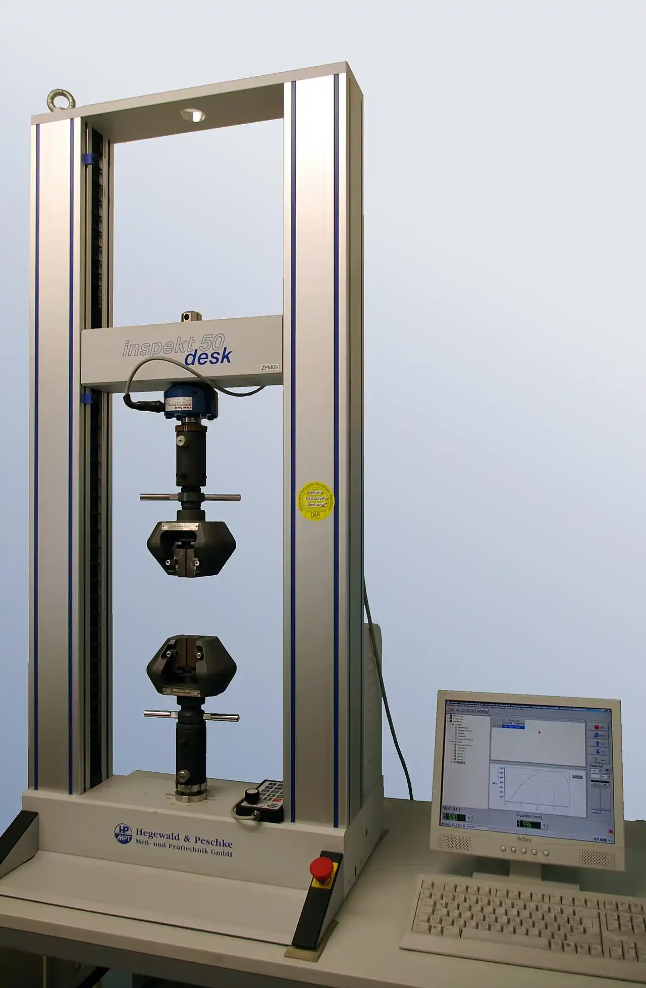 Hegewald & Peschke inspekt 50 desk twin-column electromechanical tensile testing machine with two wedge grips holding a specimen and a control computer showing the stress-strain curve