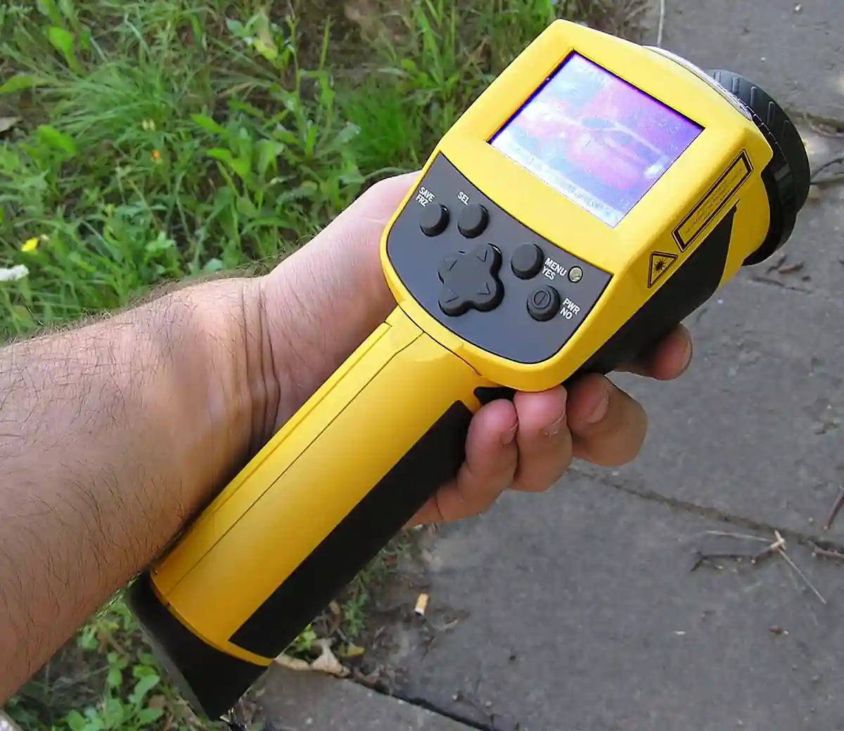 A hand holding a yellow pistol-grip thermal imager (infrared camera), its display showing a false-color thermogram of a car alongside control buttons and the infrared lens