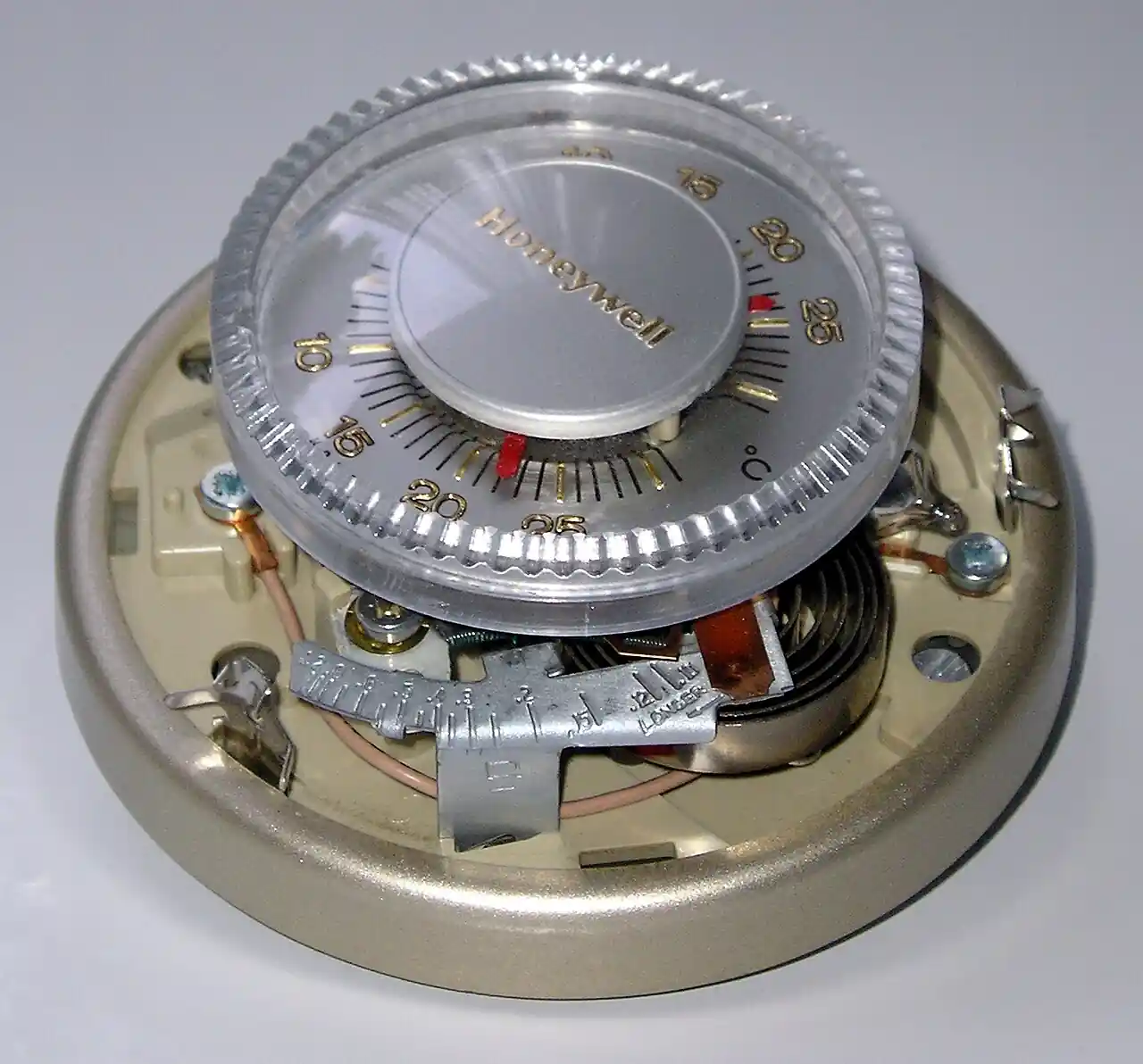 A Honeywell round room thermostat with its cover removed, exposing the Celsius-marked setpoint dial, the coiled bimetal sensing element, the heat-anticipator differential slider, and the wiring terminals